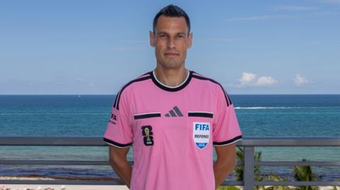 Italian referee Maurizio Mariani standing with hands behind his back wearing a bright pink FIFA Referee shirt stood in front of a white sandy beach with blue sky and sea in the background.