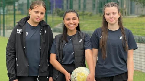 A group of three girls wearing navy blue sports clothing. Kayleigh is wearing a black rain jacket and a navy t-shirt. Jen is wearing a navy t-shirt, a black gilet with hair ties attached to it, a black skort and she is holding a netball. Tilly is wearing a navy t-shirt and black shorts. 