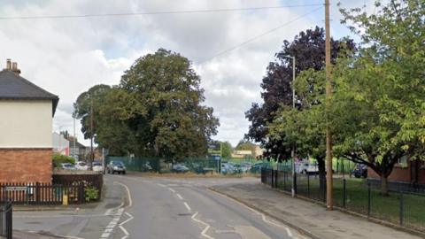 Swindon Road in Cheltenham on a cloudy day. It is a residential street, lined with trees and grass verges. Two residential streets can be seen leading off of it, as cars drive up and down. 