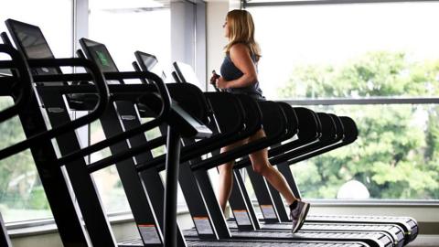 A woman wearing gym gear runs of a treadmill near large glass windows with trees outside. There are several empty treadmills next to her.