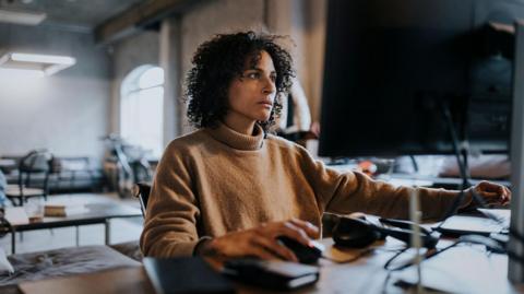 Woman with dark curly hair, wearing tan polo neck jumper, works on a computer in an office 