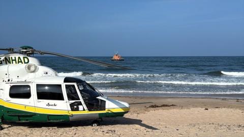 A white, yellow and green helicopter on the beach. The sea is behind it where an orange RNLI lifeboat can be seen.