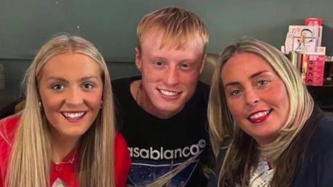 Joshua Perry is sitting in the middle of his mum and sister on a couch. They are all smiling at the camera.
