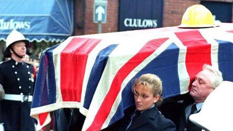 A coffin draped in a Union Jack flag and a yellow fire helmet is carried