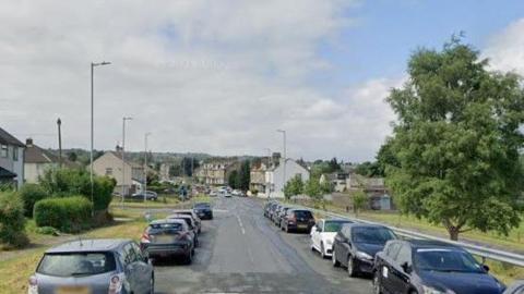 A suburban road with parked cars on both sides. A 