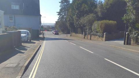 A straight road heading downhill with a new surface. Several cars are at a green traffic light at the bottom of the hill.