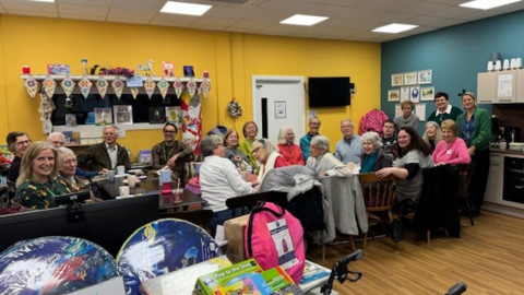 A long table with multiple people sat along it. They have turned to look at the camera and are smiling. The room has colourful walls decorated with crafts.