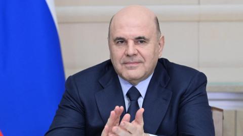 Russian Prime Minister Mikhail Mishustin claps his hands at a ceremony. The blue of a Russian flag falls behind his left shoulder.