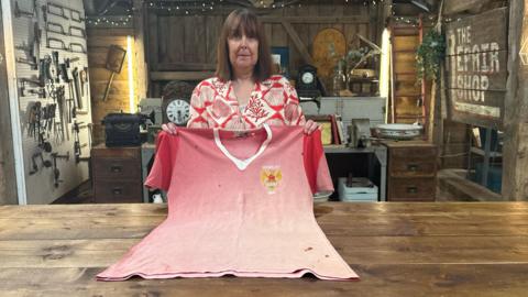 Woman with a brown bobbed haircut holds a very faded red Manchester United football shirt in front of her for the BBC's The Repair Shop programme.