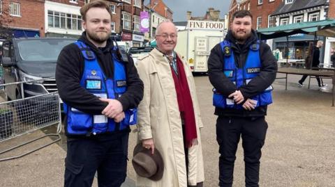 PCC Rupert Matthews standing between two blue uniformed security guards