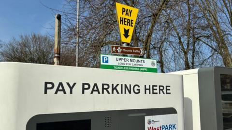 Two car park machines in a car park, with separate signs above and trees in the distance. 