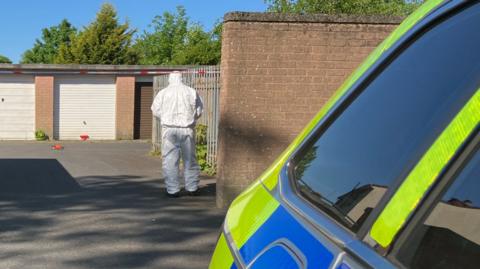 A forensic officer in a white suits walks towards a row of garages with his back to the camera. There is a white and red police tape blocking of entry to the garages which have white and brown roller doors. There is a brown brick wall next to the forensic officer. Beside the camera is a yellow and blue police car.