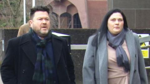 Matthew Smith and Theresa Smith walking up to Glasgow Sheriff Court. Matthew has short dark hair and a beard and is wearing a black coat and scarf. Theresa has long dare hair and is wearing a grey coat and scarf