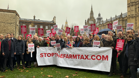 Protesting farmers gather at Westminster behind a banner reading "stopthefarmtax.com" 