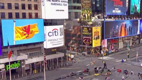 A view over Times Square in New York. There are high buildings with screens on them showing different ads. One screen is showing an image of the Somerset flag.