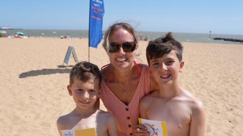 Two young boys stand either side of their mum. She is wearing a red gingham, buttn down dress and is wearing sunglasses .They are standing on a sany beach with a flag and people playing in the background.