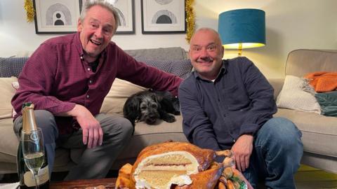 Paul Whitehouse and Bob Mortimer are grinning as they sit in a lounge. The roast turkey cake is on a table in front of them and it has been carved to reveal a Victoria sponge cake. There is also a bottle of champagne with a glass. There is a small black dog lying on the sofa next to Whitehouse and tinsel above the picture behind them.