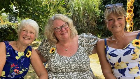 Three women sitting close together, one has short white hair, another blonde wavy hair and another with her brunette hair tied up with sunglasses on the top. They are all wearing summer dresses and have a sunflower broach attached to the neckline of the dresses. 