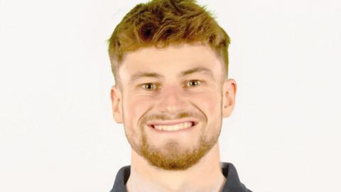 Joe Shingler, wearing a Team UK grey polo shirt. He has short ginger hair and beard and is smiling at the camera against a white backdrop 