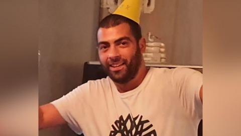 Brian Rogers smiles at the camera. He is wearing a white T-shirt with a singular pattern in a dark colour on the front of the T-shirt. He has a yellow party hat on his head.