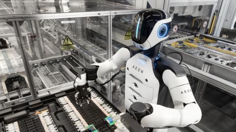 The Aeon robot at work in the factory. It has a white torso with black shoulders. Its black plastic face has a vertical line of blue light.