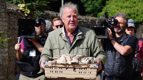 Jeremy Clarkson carrying mushrooms at the opening of his new pub, The Farmer's Dog. He is wearing a green coat, and is being followed by two camera men.