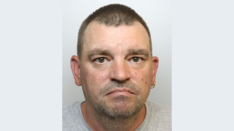 A police mugshot of Matthew McKenzie, wearing a grey t-shirt and standing against a plain grey background. He has short brown hair, brown eyes, a silver hoop earring in his left ear and slight stubble. He is looking at the camera with a sad expression.