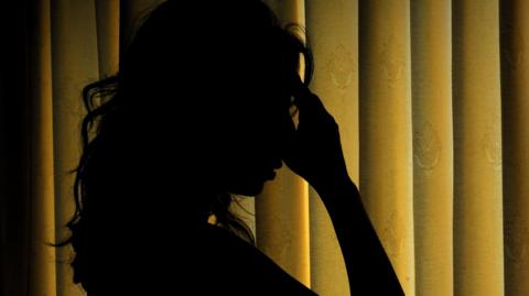 The silhouetted woman with long hair and her hand to her head in front of curtains