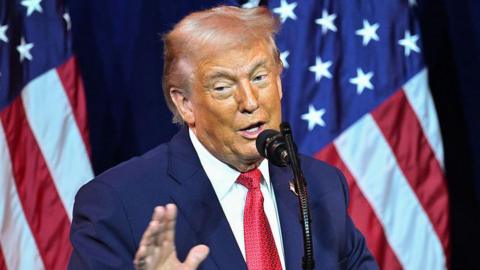Donald Trump mid-speech. He's in a blue suit, white shirt and red tie, two US flags on poles behind him