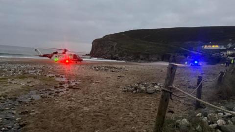 A white and red helicopter with a bright red light sits on a sandy beach, strewn with stones at low tide. In the background a bright blue light from an emergency services vehicle at the edge of the beach, below cliffs and a headland. A rope fences off some maram grass in the foreground.