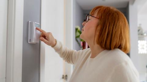 A woman with red hair cut in a bob is wearing glasses and a white long-sleeve top. The photo captures her from her left hand side in profile as she raises her right hand to adjust a thermostat on the wall of her house. The wall is painted grey, beside it is a white door and doorframe.