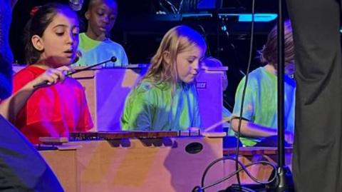 Children are seated on a stage playing glockenspiels - two girls, one in a red T-shirt, the other in a green one, sit in front of a boy, who is also wearing a green T-shirt 