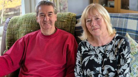 Kenneth and Carol are sitting on a sofa, draped with blankets. Kenneth is wearing a red sweater and smiling at the camera. Carol has shoulder-length blonde hair and a blue and white floral top. 