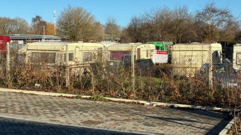 Around seven vehicles parked inside a meanwhile site, behind a wire mesh fence. 