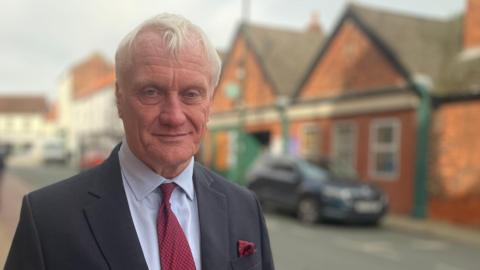 Graham Stuart - a man with short white hair wears a smart dark suit, pale blue shirt and red tie. He is standing in a street. The background is out of focus and shows red-brick buildings, cars and a road. 