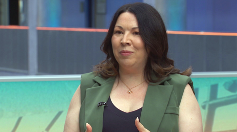 Monica Lennon has shoulder-length, dark hair. She is wearing a black vest with a green suit-style waistcoat over it. She is mid chat
