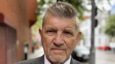 Shows a man with a beard. He wearing a grey jacket with a white shirt and blue striped tie. 