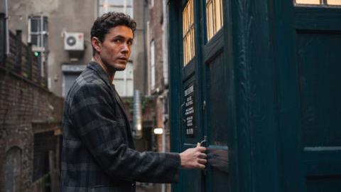 Doctor Who character Conrad, with a print blazer and curly brown hair, opens door to Tardis