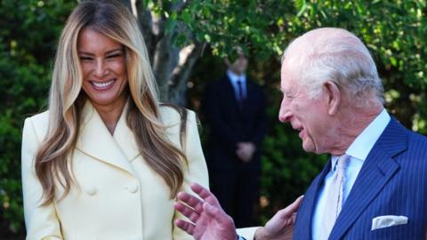Melania Trump and King Charles laughing together at the White House. Melania Trump is wearing a cream suit and King Charles is wearing a blue suit