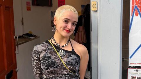 A woman with really short blonde hair, wearing an off-the-shoulder top standing outside a fashion studio