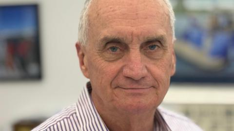 Paul Callingham is looking at the camera with a small smile. He has short white hair and is wearing a striped shirt.