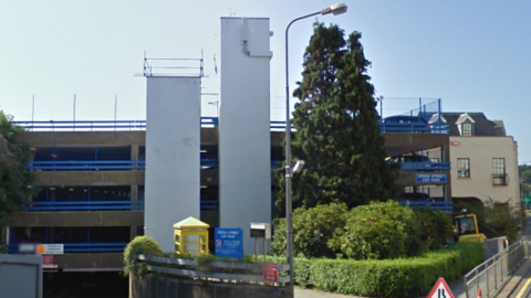 Four levels of a multi-storey car park are in the image, with blue rails along the edge of each floor. There are two floor to ceiling white towers besides the building.