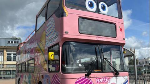 A pink open top double decker bus with Hattie written on the front and big eye stickers on the top windows. The bus is colourfully decorated. It is parked in Scunthorpe in front of a bus station building with St John's Church in the distance to the right.