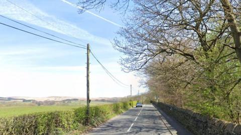 Street view image of Horrobin Lane, Rivington, a country lane with a field to the left and trees to the right. The sun is shining and the sky is blue with aeroplane trails across it