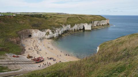 A coastal cove with a sandy beach nestled between tall white cliffs. The scene appears to be taken from an elevated grassy viewpoint looking down into the bay. The beach sits in a curved, sheltered bay formed by steep chalk cliffs. A few people or objects appear to be floating or paddling in the water near the cliffs.