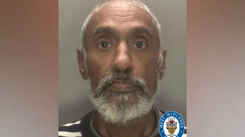 A police mugshot of a man with a grey and white beard.