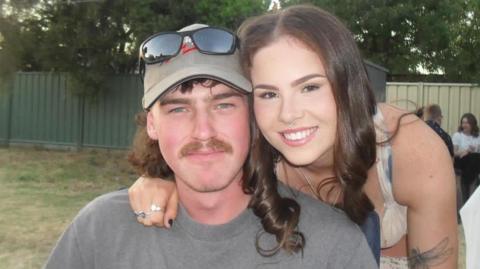 Justyce Crossman with her arm around her fiancee David Robinson. Both are in their early twenties. Justyce has long, brown hair and brown eyes. She has a septum piercing and a tattoo on one of her arms of a dragonfly. David has a brown mullet and moustache and blue eyes. He wears a baseball cap with sunglasses perched on top.