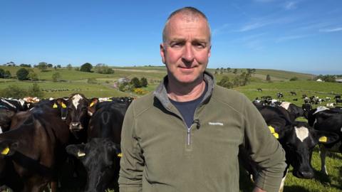 Robert Craig, a man with balding, short hair, wearing a green fleece and navy t-shirt, looks at the camera. A herd of black and brown cows are behind him in rolling green fields. It is a sunny day with blue skies. 