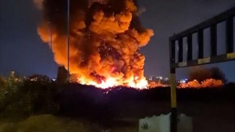 A large fire rising up, showing lots of flames and smoke. There are bushes in front of the flames and the sky is dark.