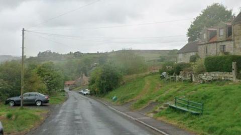 A steep road in a country village. Terraced cottages sit on one side of the road behind a grassy bank, which also features a green sitting bench. A car is parked behind a telegraph pole on the other side of the road.